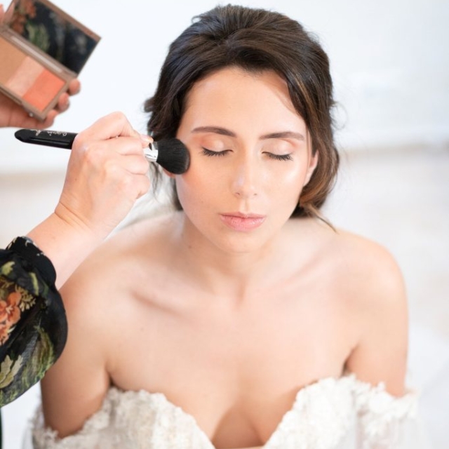 A bride in an off-shoulder wedding dress sits with eyes closed while makeup is applied to her cheek with a brush.