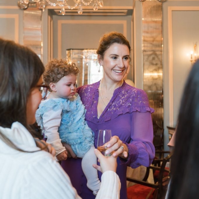 A woman in a purple dress holds a baby in a blue outfit, smiling and talking with others as light from chandeliers glimmers across the ornate décor and patterned lanes of the elegant room.