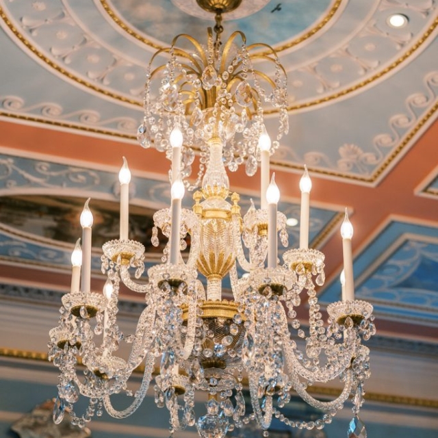 An ornate crystal chandelier with lit candles hangs from an intricately decorated ceiling with pastel colors and gold accents, casting a soft glow over the elegant halls of The Lanes.