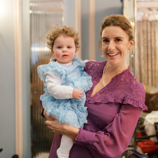 A woman in a purple dress holds a baby in a blue ruffled outfit while smiling at the camera, capturing a joyful moment together indoors along The Lanes.