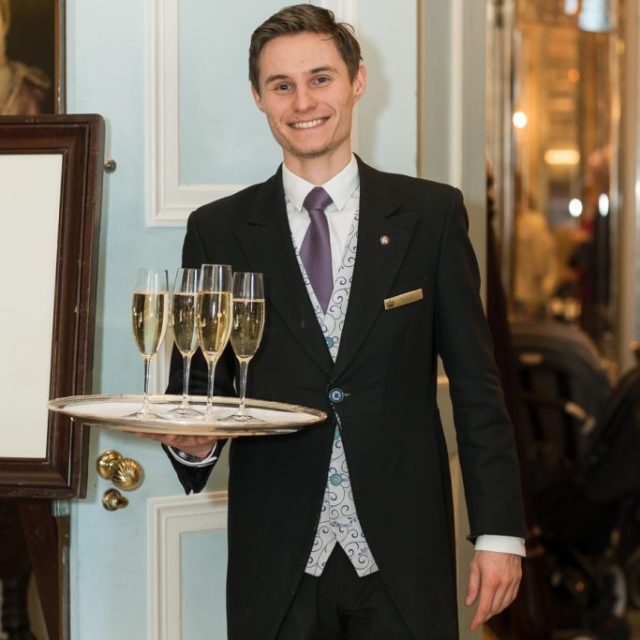 A smiling waiter in formal attire holds a tray with four glasses of champagne in an elegant indoor setting at The Lanes.