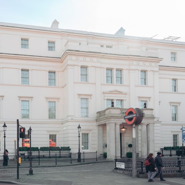 White neoclassical building with multiple windows stands behind an entrance to the London Underground near The Lanes; several people are walking along the sidewalk in front.