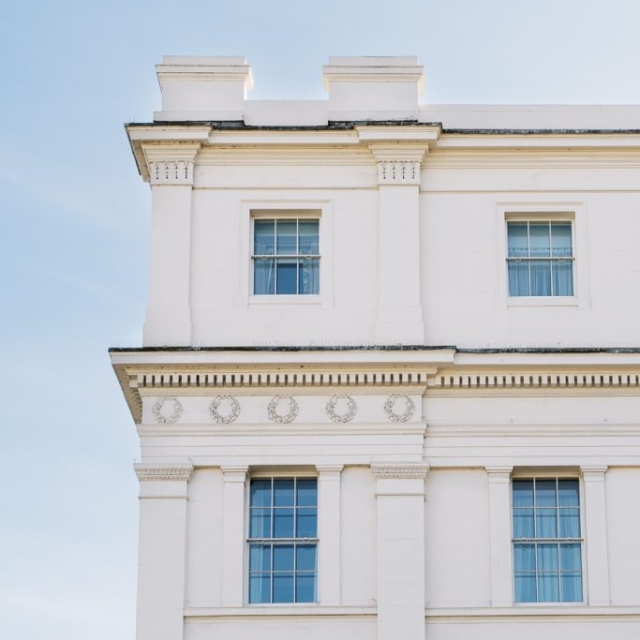 White neoclassical building facade along the lanes, featuring three visible windows, decorative molding, and columns against a clear blue sky.