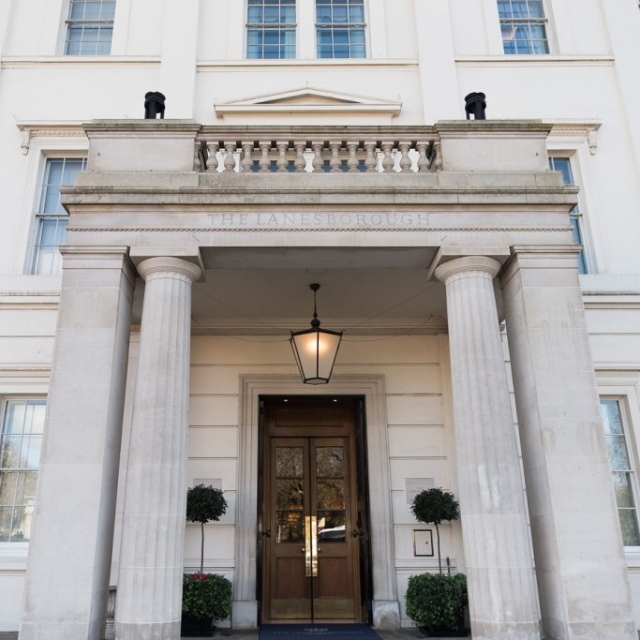 Elegant white building with four large columns at the entrance, glass double doors, potted plants flanking the lanes leading up to the entryway, and a lantern hanging above the doorway.