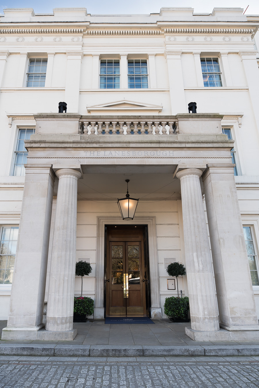 A white neoclassical building facade with four large columns frames the entrance of the Lanesborough Hotel in London, where two potted plants stand ready to welcome guests for a First Birthday Celebration.
