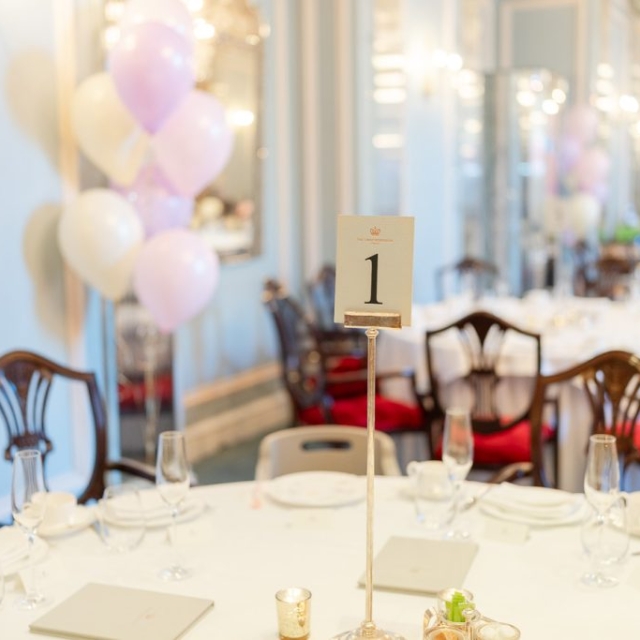 A banquet table labeled "1" with empty plates and glasses in a decorated dining room; pastel balloons float in the background, creating a festive atmosphere at The Lanes.
