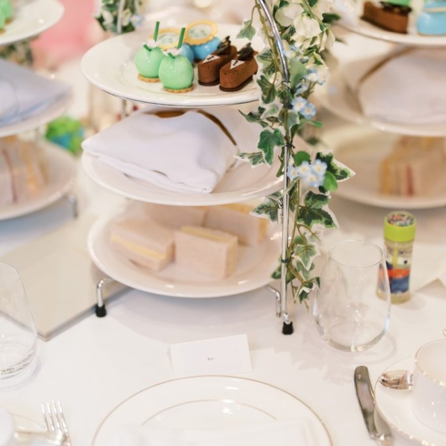 A three-tiered stand with assorted pastries and sandwiches, decorated with green ivy, sits on a white tablecloth set near plates, glasses, and cutlery—perfect for elegant gatherings in tea lanes.