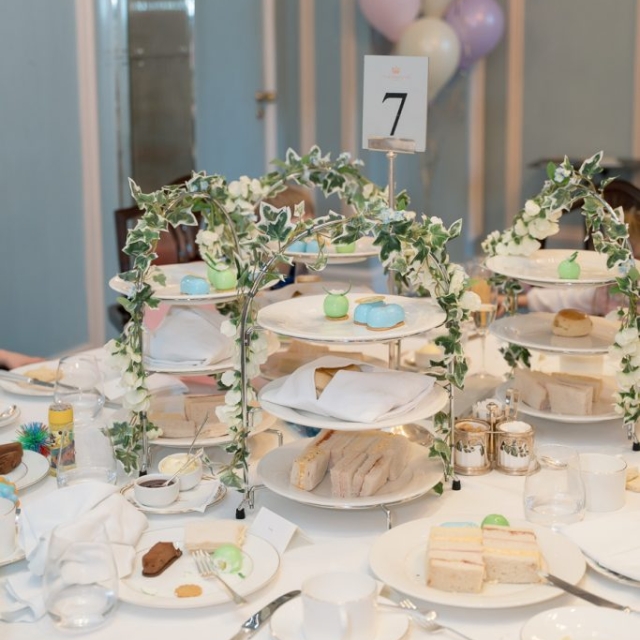 A round table at The Lanes set for afternoon tea with tiered trays of sandwiches, pastries, and desserts; table number 7 is displayed, with pastel balloons in the background.