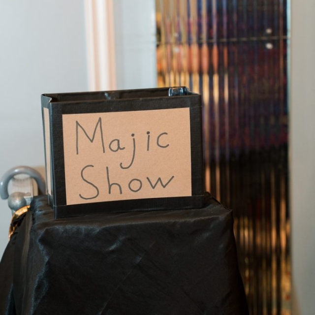 A hand-drawn sign reading "Majic Show" is displayed on a box draped with a black cloth, set in The Lanes against a metallic, reflective background.