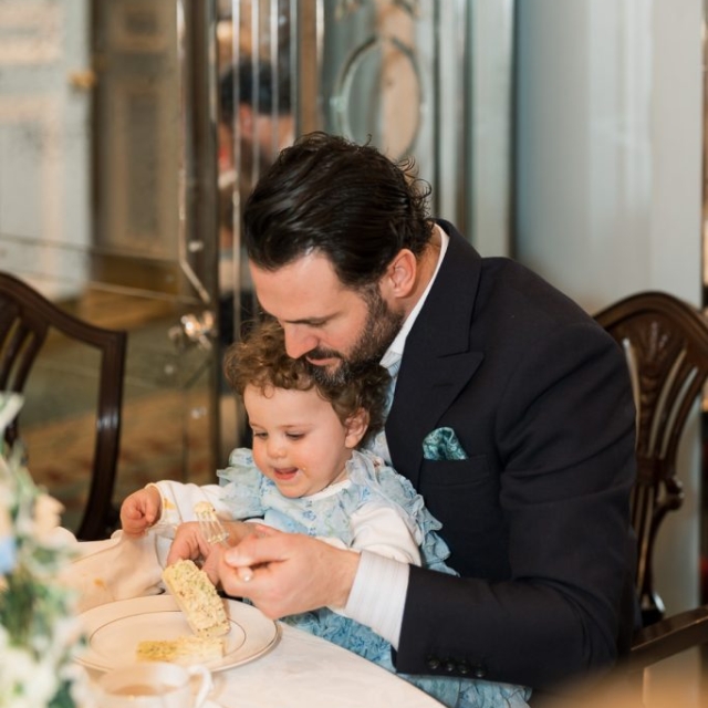 A man in a suit helps a young child eat at a dining table at The Lanes, both seated closely together and smiling.