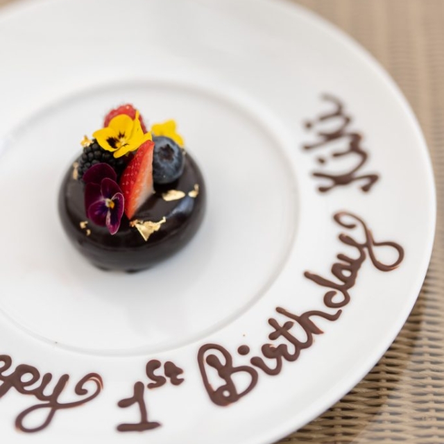 A small chocolate dessert from The Lanes, adorned with berries, edible flowers, and gold leaf, sits on a plate with "Happy 1st Birthday Kiki" written in chocolate.