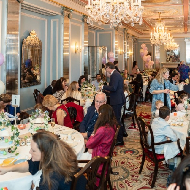 Guests seated at round tables lining subtle lanes in an elegant, chandelier-lit room decorated with balloons, engaged in conversation and dining during a formal event or celebration.