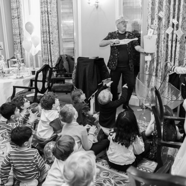 A magician performs a trick for a group of children seated on the floor at The Lanes, with one child assisting and holding an umbrella catching falling cards in a decorated room.