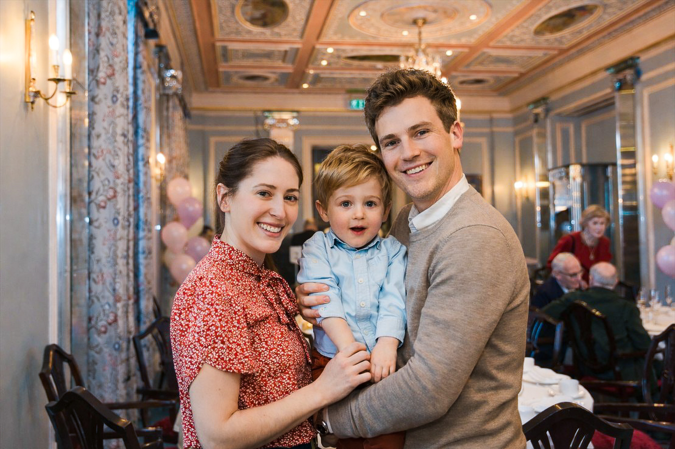 A smiling couple holds a young boy in an elegant room decorated with pink balloons; people are seated at tables in the background.