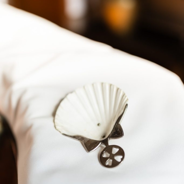 A white seashell-shaped dish with a metal holder and a round keychain labeled "Brouillon auto" rests on a white fabric surface.