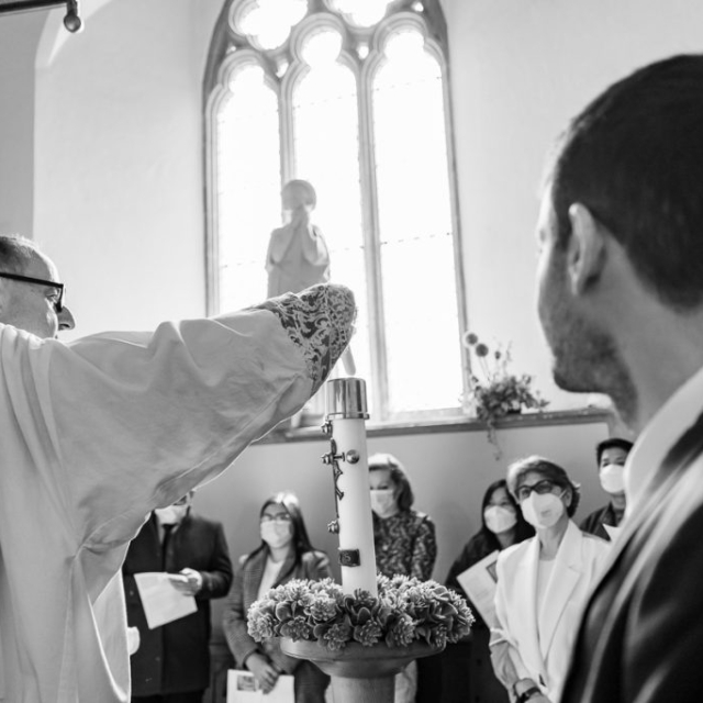 A priest lights a large candle during a church ceremony as a group of people, some wearing masks, and others holding Brouillon auto papers, watch attentively.