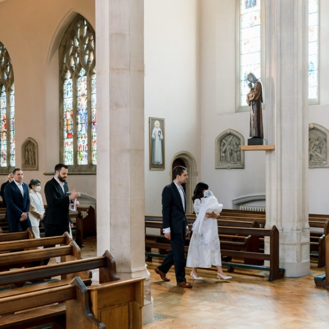 People dressed formally walk down the aisle of a church with wooden pews, stained glass windows, and religious statues visible.