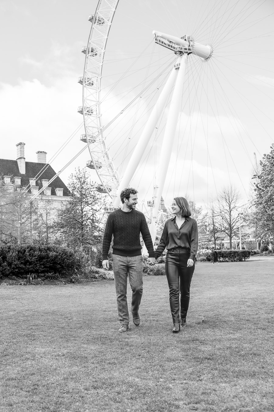 Un homme et une femme marchent ensemble sur l'herbe devant le London Eye, se tenant par la main et se regardant l'un l'autre. La scène est en noir et blanc.