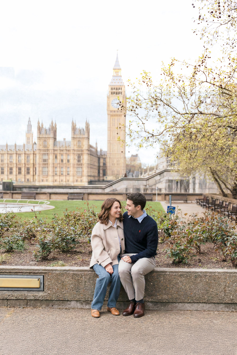 A couple is sitting on a concrete wall, smiling at each other, with the Palace of Westminster and Big Ben in the background on a cloudy day.
