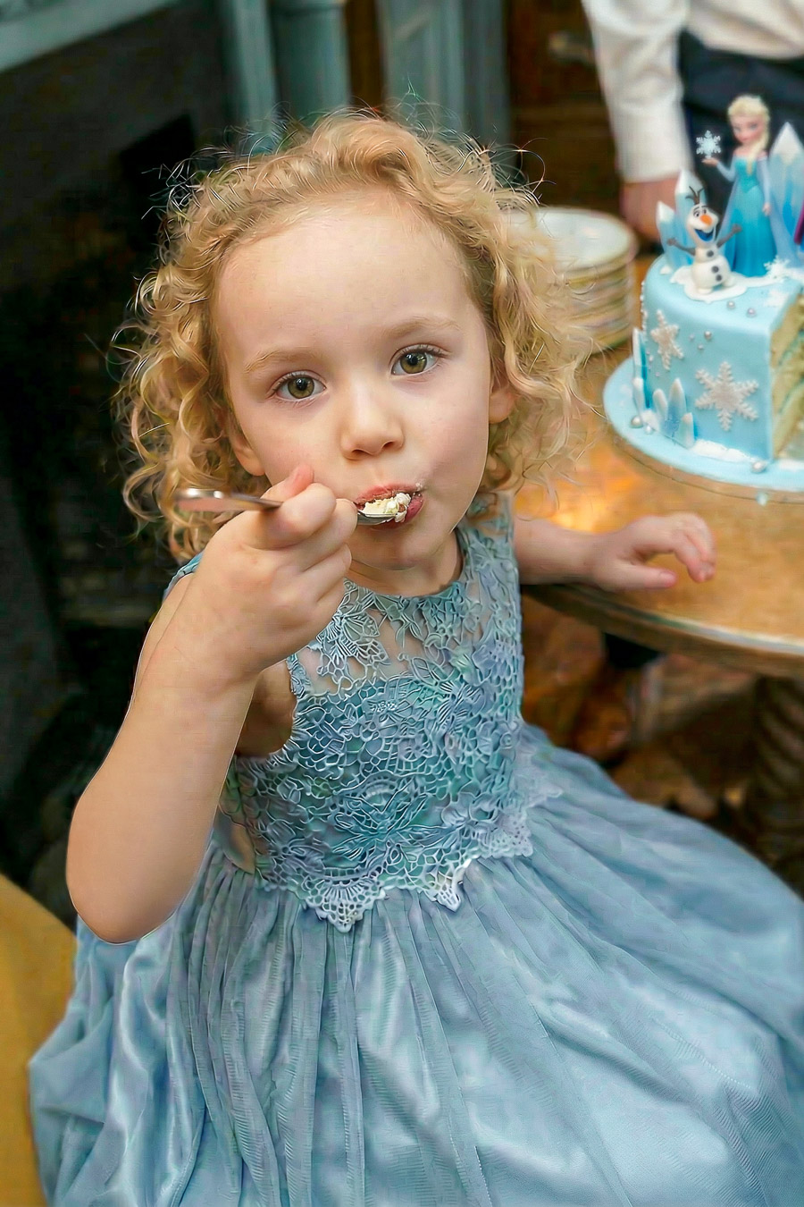 A young child in a blue lace dress eats cake with a fork, sitting at a table with a blue Frozen-themed cake in the background.