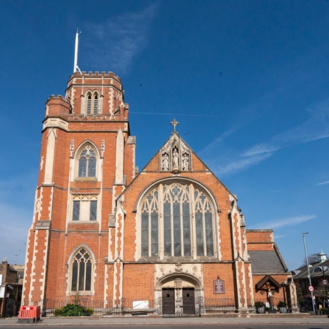 A large brick church with a tall tower and arched windows stands under a blue sky. A small Brouillon auto sign is visible on the fence, and nearby buildings are in the background.