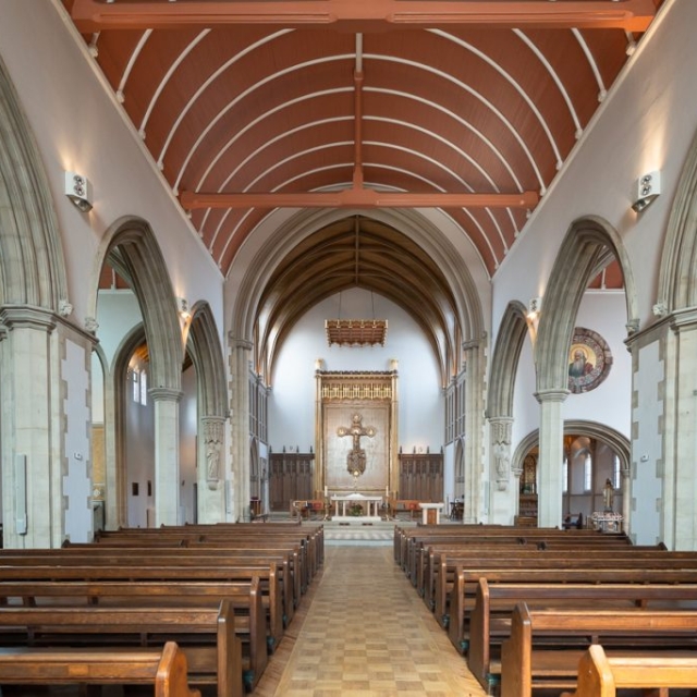 Interior of a church with wooden pews, arched columns, stained glass windows, and a Brouillon auto at the altar featuring a large cross at the far end.