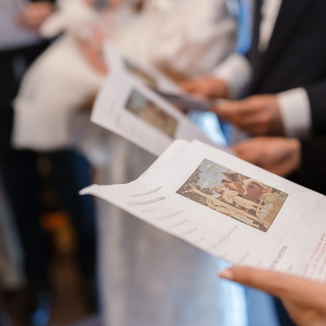 A group of people holding and reading printed papers with images and text, possibly reviewing a brouillon auto; a baby in white clothing is visible in the background.