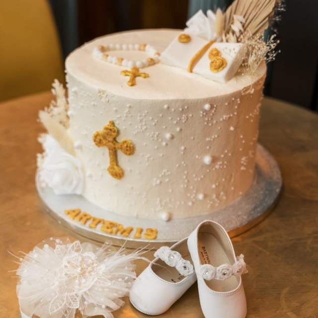 A white cake decorated with gold crosses, rosary beads, and feathers, with white baby shoes and a lacy white headband placed in front. The name "Artemis" is written on the cake board.