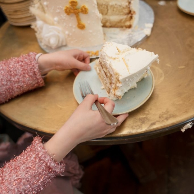 A child in a pink outfit sits at a table, holding a fork and plate with a slice of layered white cake decorated with a gold cross. The rest of the cake is on the table.