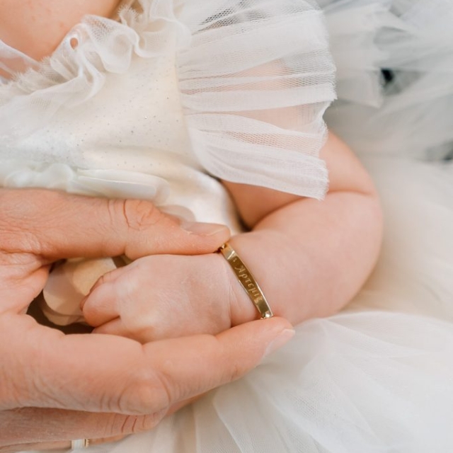An adult hand gently holds a baby's hand. The baby wears a white dress with tulle and a gold bracelet with engraved letters.