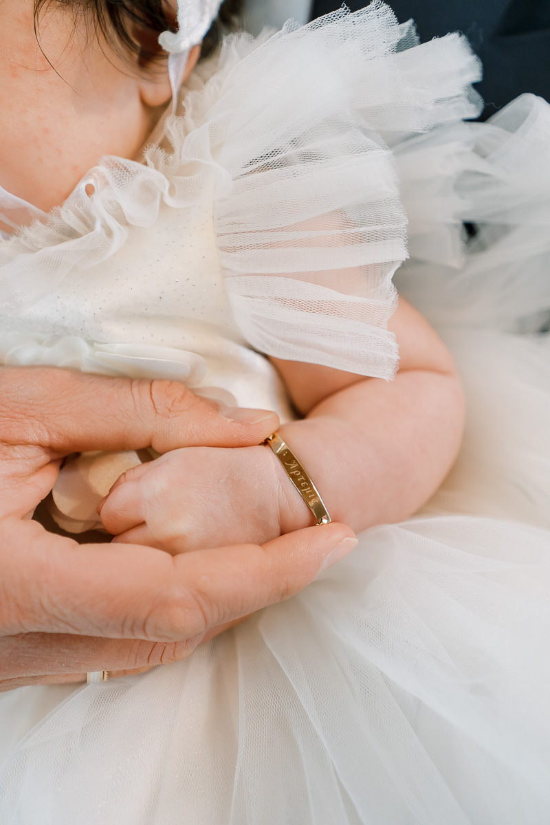 An adult hand gently holds a baby's hand. The baby wears a white dress with tulle and a gold bracelet with engraved letters.