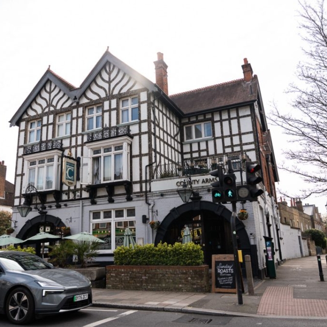 A black and white timber-framed pub called "The County Arms" stands at a street corner, with outdoor seating, a car passing by, and a Brouillon sign near the pedestrian crossing in view.