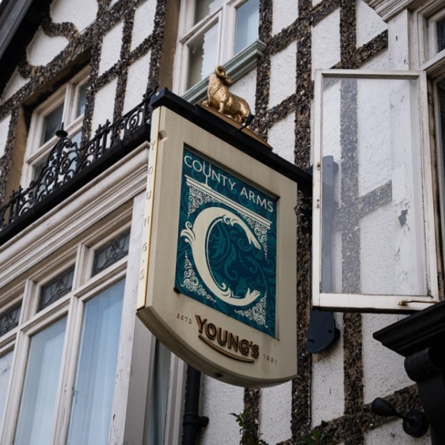 A pub sign reading "County Arms" and "Young's" hangs outside a Tudor-style building, topped with a gold ram statue. An open window is nearby.