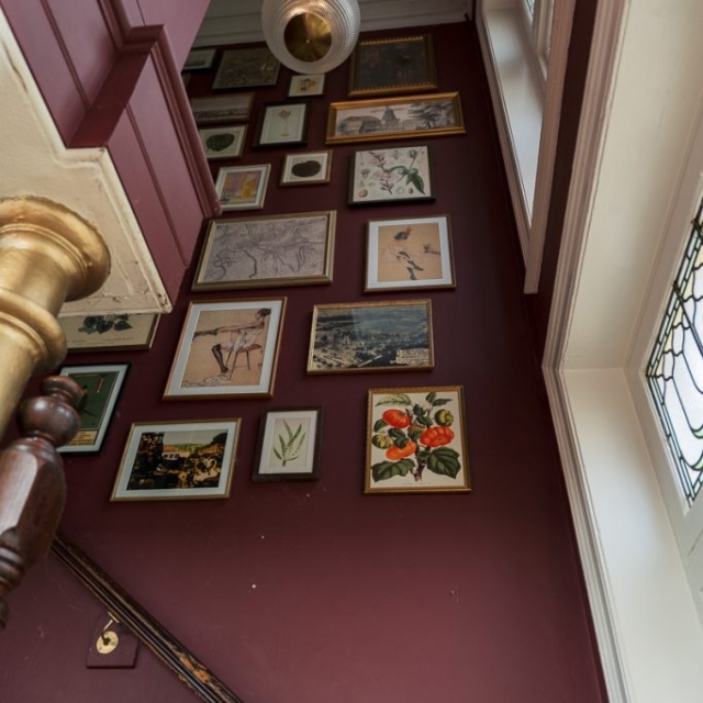 A maroon stairwell with framed artwork on the wall, a brass handrail, a hanging light fixture, and a sign pointing to "Trinity Bar.
