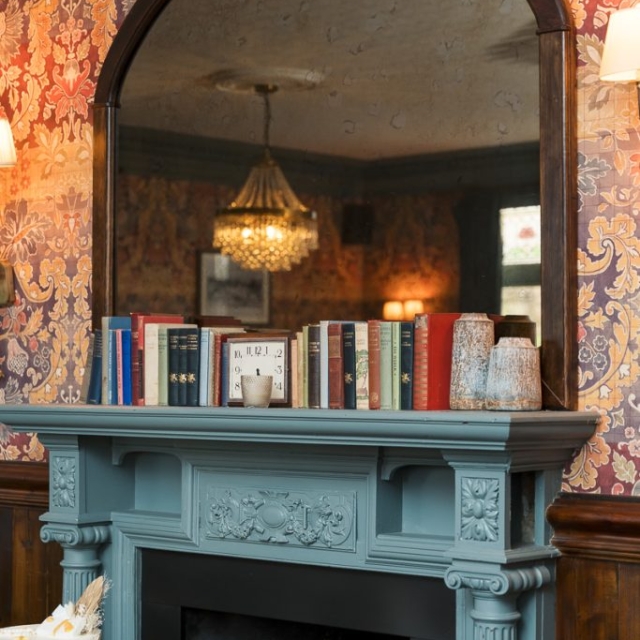 A blue fireplace mantel with books, candles, and framed art sits below a large arched mirror, reflecting a chandelier in a room with floral wallpaper.