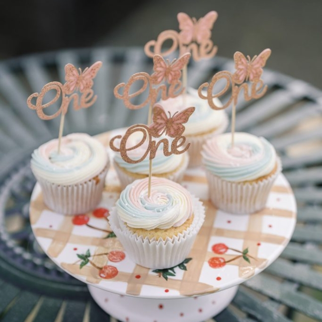 Six pastel-frosted cupcakes on a cake stand, each topped with a butterfly and "One" decoration, displayed on an outdoor metal table.