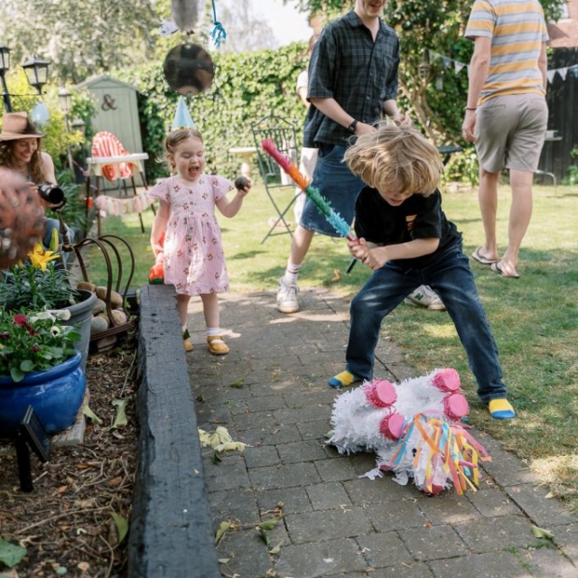 A young boy hits a unicorn piñata with a stick while a toddler in a dress watches at an outdoor party with adults nearby in a garden.