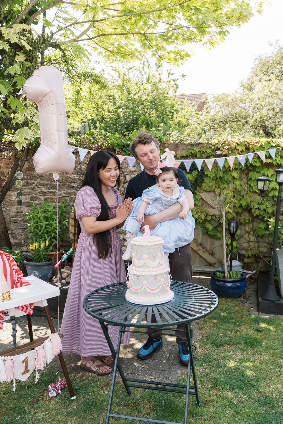 A woman, man, and baby stand outdoors behind a two-tiered cake on a table, with a large number one balloon and party decorations—capturing the joy of first birthday party photography perfect for Family Photographer London or North Northamptonshire.