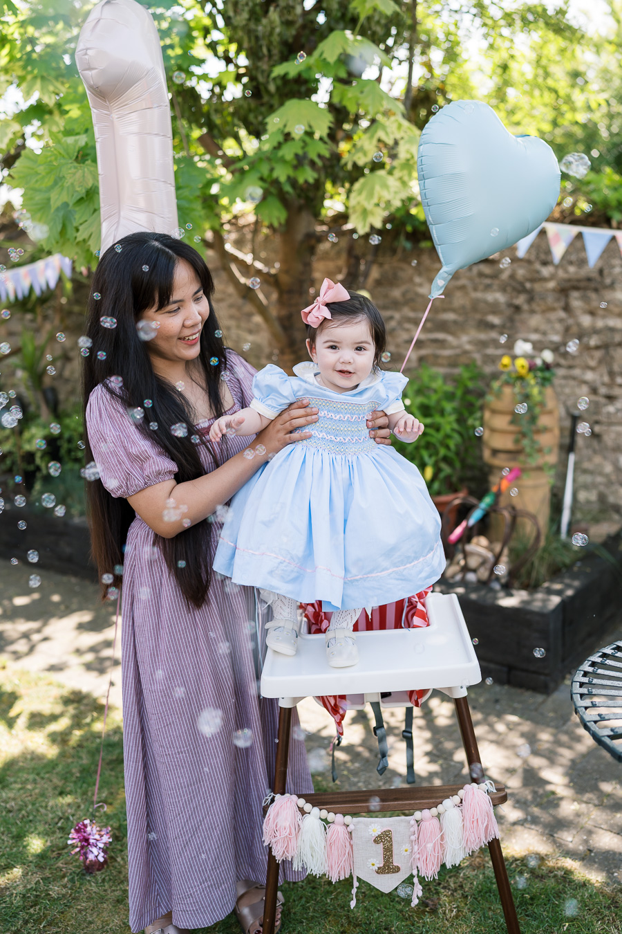 A woman stands beside a toddler in a high chair outdoors, holding a blue heart balloon. The high chair features a "1" banner, and bubbles float around them.