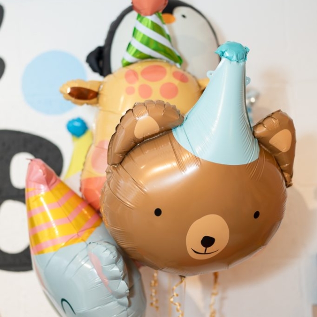 A group of animal-shaped balloons, including a bear, elephant, and giraffe, all wearing party hats, are tied together against a white wall background.