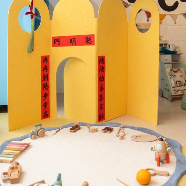 A children's play area with a yellow screen featuring red and black Chinese calligraphy, surrounded by wooden toys on a round white rug.