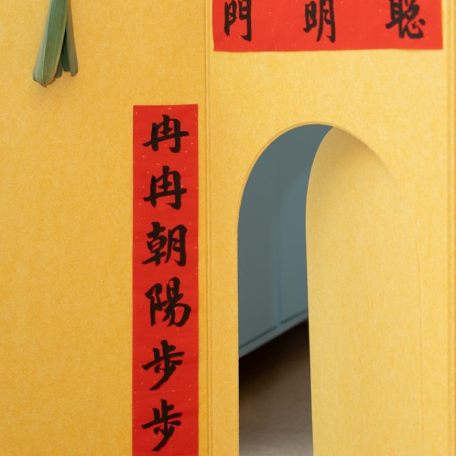 A yellow wall with red Chinese calligraphy banners and a green plant stem hanging through a circular cutout above an arched doorway.