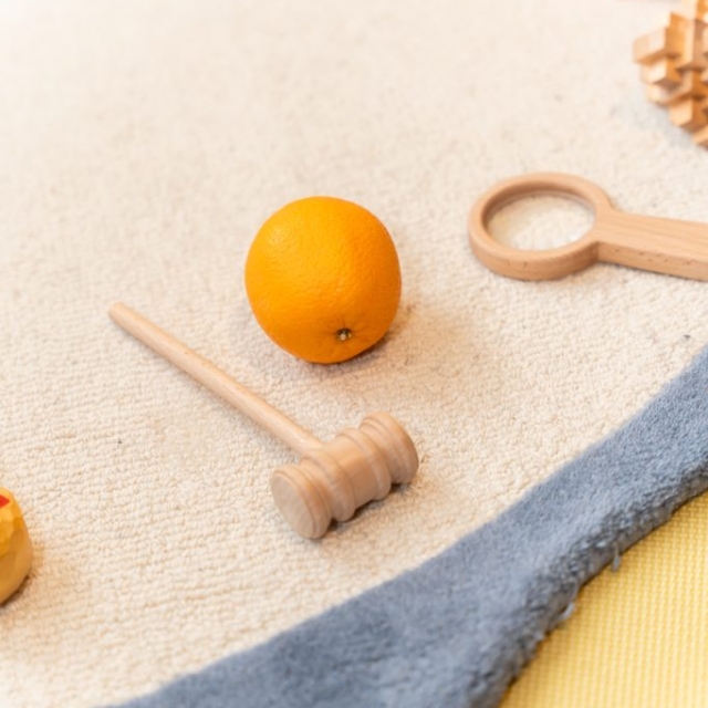 An orange, a wooden mallet, and a wooden ring toy are placed on a light-colored carpet with a blue edge.