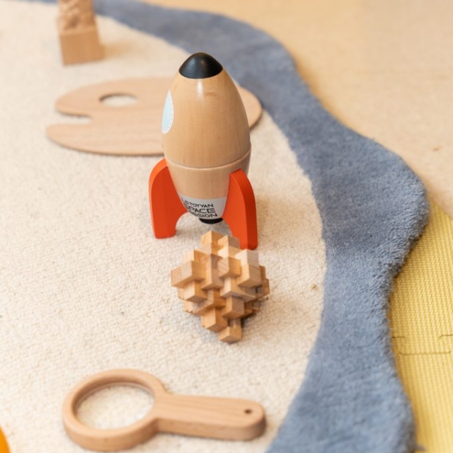 Wooden toy rocket, a puzzle, a magnifying glass, and other wooden toys are arranged on a light carpet next to a gray and yellow border.