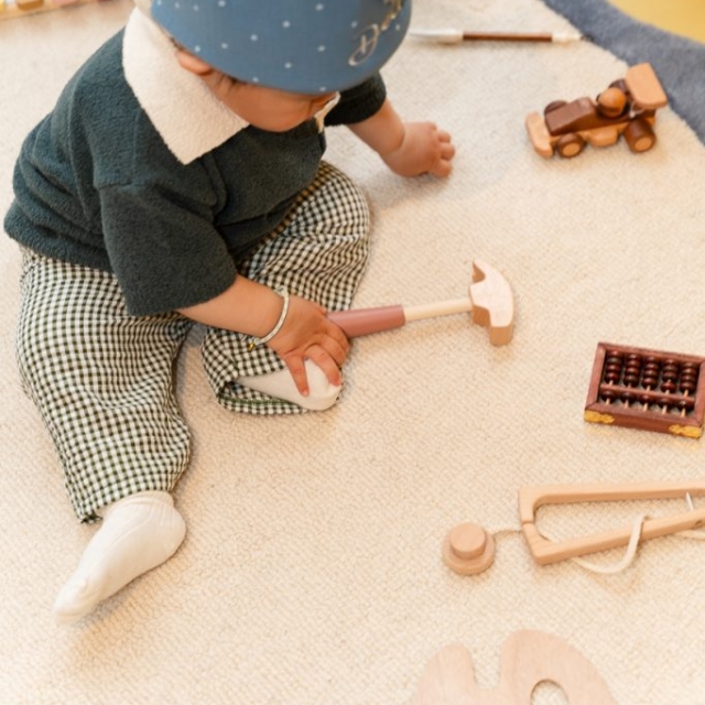 A baby wearing a blue crown sits on a rug, holding a wooden hammer, surrounded by wooden toys including a train, abacus, and xylophone.
