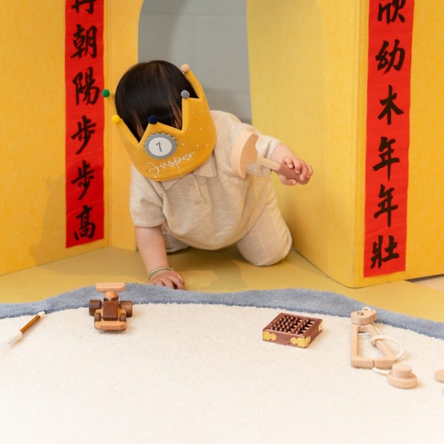 A toddler wearing a yellow crown crawls through a yellow play structure decorated with red banners, surrounded by wooden toys on a white floor.