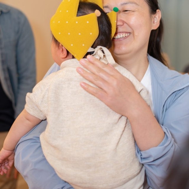 Woman holding a baby wearing a yellow crown with colorful tips; both are smiling indoors in a well-lit room.