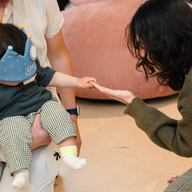 A woman reaches out to hold hands with a seated baby wearing a blue crown, who is sitting on an adult’s lap indoors.