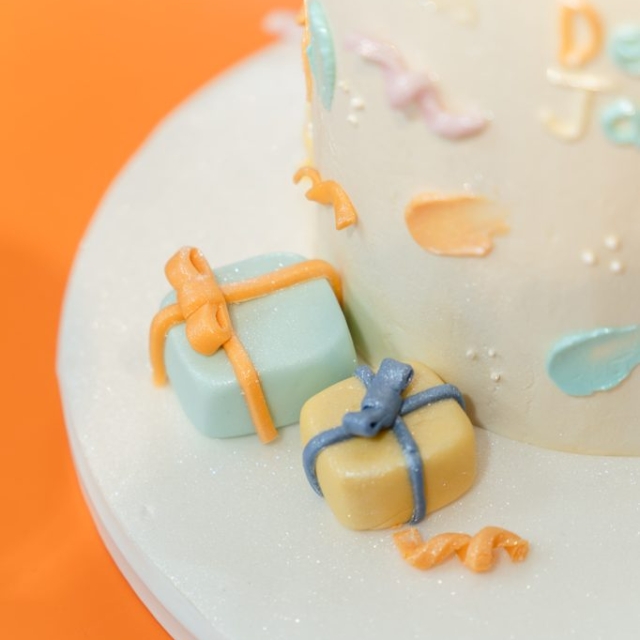 Close-up of a white frosted cake decorated with pastel-colored fondant presents and ribbon details, set on a white cake board with an orange background.