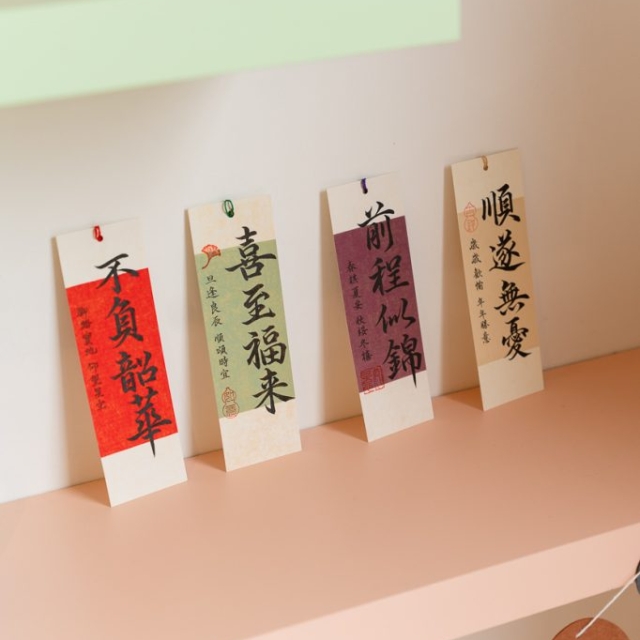 Four vertical cards with Chinese calligraphy are displayed upright on a peach-colored shelf, each card clipped with a small colored paperclip.